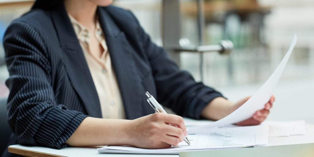 Close up of young businesswoman writing in document while working at cafe table in office building setting, copy space