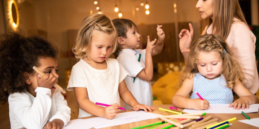 multicultural kids drawing in kindergarten, educator looking at gesturing boy