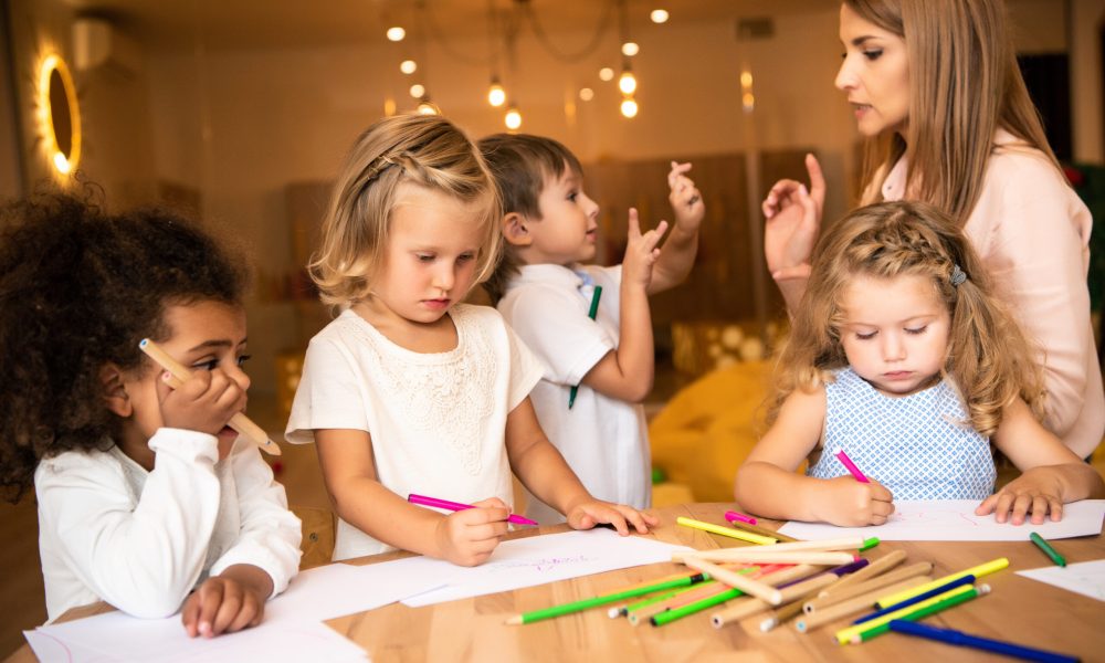 multicultural kids drawing in kindergarten, educator looking at gesturing boy